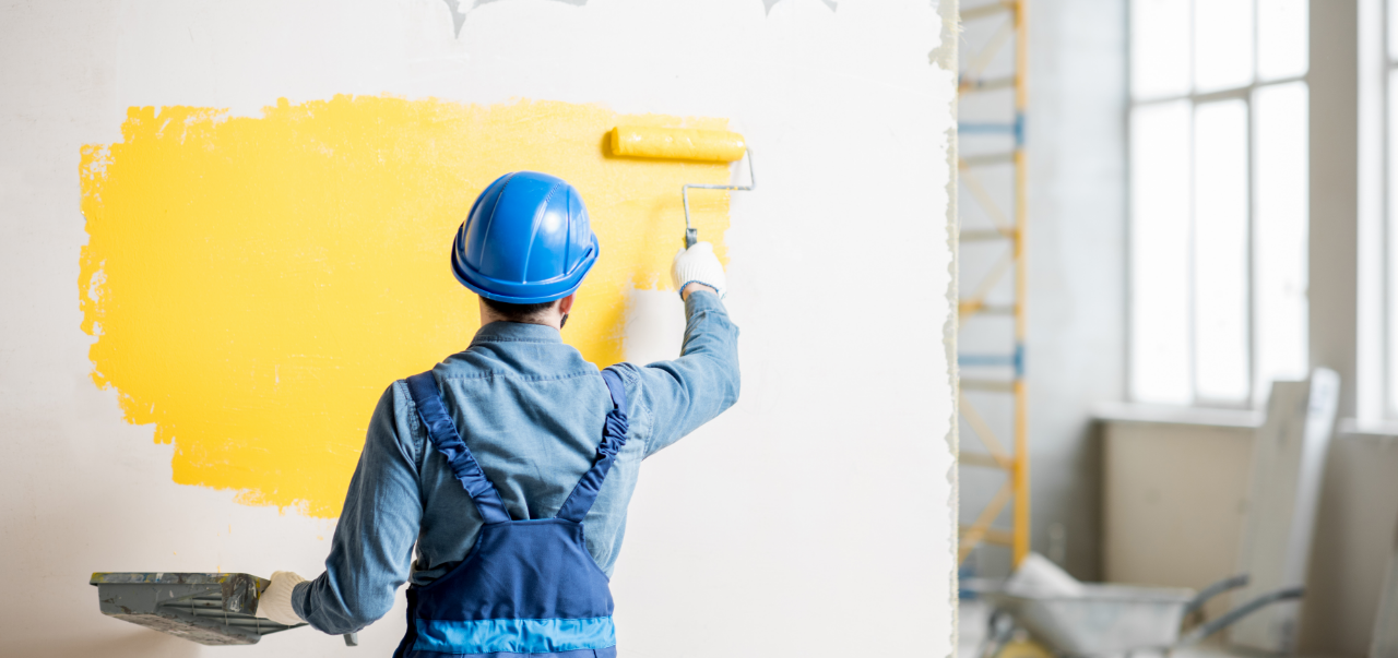 Workman in uniform painting wall with yellow paint at the construction site indoors.
